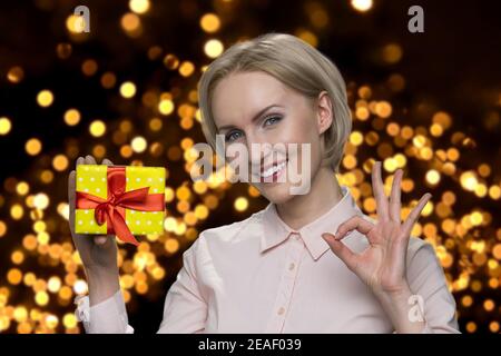 Ältere Frau hält Geschenkbox und zeigt okay Geste. Stockfoto