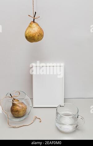 Modernes Stillleben mit weißem leeren Bilderrahmen. Sommerkomposition mit Birnen und einem Glas Wasser auf einem hellen Tisch. Layout. Stockfoto