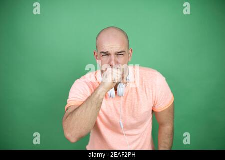 Junge Glatze Mann mit Kopfhörer über grünem Hintergrund mit seinem Hand an den Mund husten Stockfoto