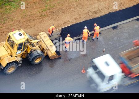 Asphalt wird mit Schaufeln aus dem Eimer eines Bulldozer während der Straßenarbeiten gelegt, Draufsicht Stockfoto