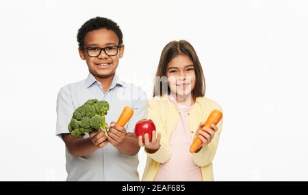 Latino Mädchen und afroamerikanischen Jungen halten Brokkoli, Karotten und einen Apfel. Vorteile von Gemüse und Obst für Kinder Stockfoto