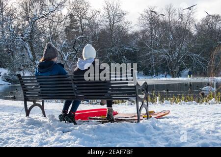 Glasgow, Schottland, Großbritannien. 9th. Februar 2021. UK Wetter: Zwei Mädchen sitzen auf einer Bank neben dem gefrorenen Teich im Queen's Park nach starkem Schneefall von Storm Darcy. Kredit: Skully/Alamy Live Nachrichten Stockfoto