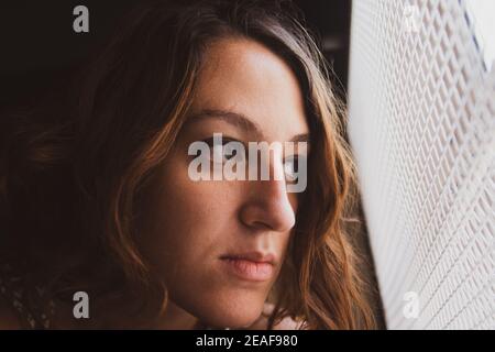 Traurige junge Frau, die aus dem Fenster schaute. Einengung in Zeiten des Coronavirus. Wunsch nach Ausgehen Stockfoto