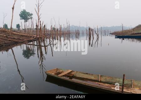 Altes Holzboot in kleiner Bucht des Nam Theun Flusses. Tote trockene Bäume spiegeln sich im Wasser. Traditionelle ländliche Landschaft Blick in Laos. Thalang, Thakhek, Laos Stockfoto