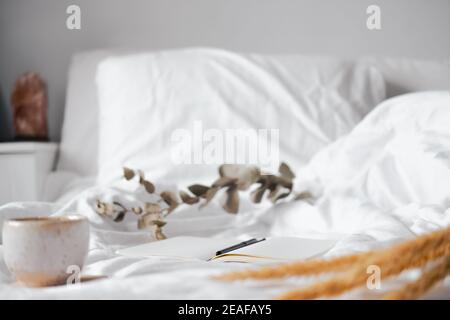 Offener Notizblock mit leeren Seiten in Textil-weißem Hintergrund, getrockneten Blumen und einer Tasse Tee. Ruhig und achtsam zu Hause leben, im Bett bleiben, Hand schreiben Stockfoto