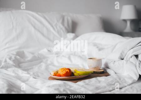 Tablett mit Obst und einer Tasse Tee in weißem sauberen Bett. Achtsam und ruhig leben, langsames Leben, Bett in Frühstück Konzept, einfache Freuden zu Hause Stockfoto