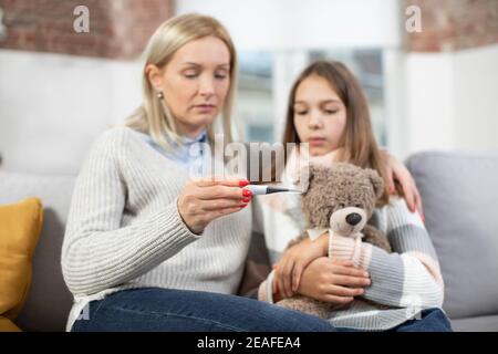 Reife blonde Frau, die nach der Messung die Temperatur ihrer kranken Tochter auf dem Thermometer überprüft. Trauriges krankes Mädchen, hält Teddybär, schaut auf Thermometer Stockfoto