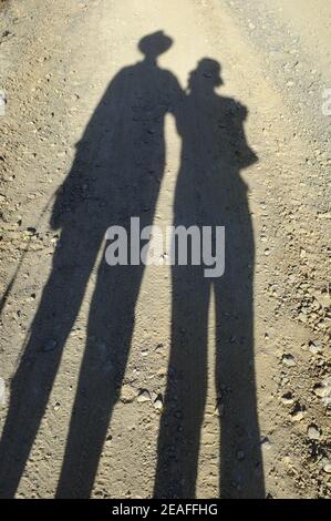 Lange Schatten eines Mannes und einer Frau, die an einem sonnigen Nachmittag auf einer staubigen, undurchmessenen Landstraße spazieren. Andalusien, Spanien Stockfoto