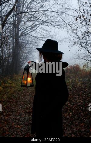 Die junge Frau geht durch den dunklen Wald, sie hält eine eiserne Laterne. Neblige Landschaft im Hintergrund. Stockfoto