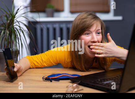 Schöne blonde Frau mit Laptop und Smartphone über Home Office Hintergrund mit Hand auf Mund. Schlafmaske auf dem Tisch, schockiert und Überraschungsgesicht Stockfoto