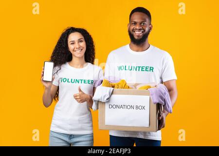 Zwei Freiwillige Zeigen Telefon-Bildschirm Holding Spende Box, Gelber Hintergrund Stockfoto