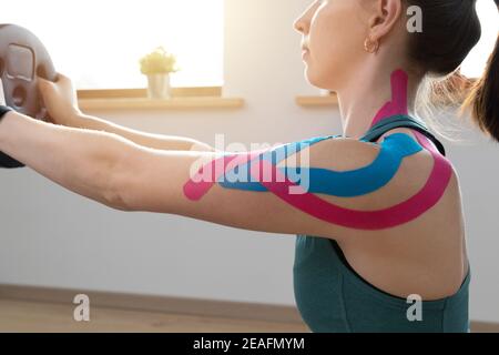 Junge, fette Frauen, die an einem sonnigen Tag zu Hause mit elastischem therapeutischen kinetischen Band auf ihrer Schulter trainieren. Kinesiologie Physikalische Therapie. Stockfoto