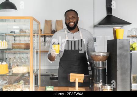 Ein freundlicher afroamerikanischer Kellner, der eine Tasse Kaffee zum Mitnehmen bestellt, ein multirassischer Barista, der Handschuhe und Schürze trägt, bedient den Gast, schaut sich den an Stockfoto