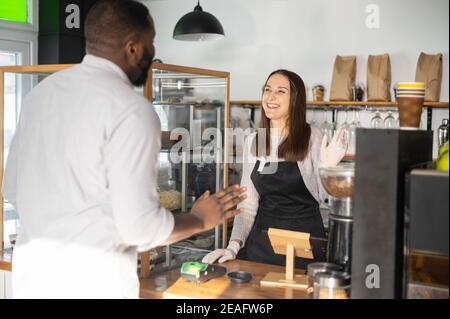 Die freundliche und hilfsbereite Kellnerin bedient einen afroamerikanischen männlichen Gast und begrüßt den Cafe-Kunden. Weibliche Bäckereibesitzerin freut sich, einen Gast zu sehen Stockfoto