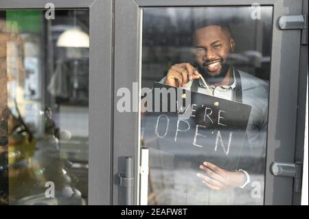 Ein fröhlicher schwarzer Kerl in einer Schürze hält in den Händen ein offenes Schild, Blick durch eine Glastür. Ein multirassischer Kellner, Manager eines kleinen Ladens Stockfoto