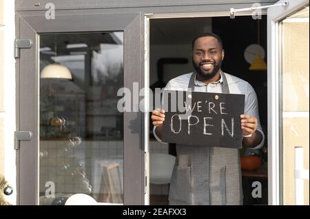 Konzept für kleine Unternehmen. Lächelnder afroamerikanischer Typ Backwaren in einer Schürze hält ein offenes Schild in seiner Hand, das außerhalb des Cafés steht. A Stockfoto