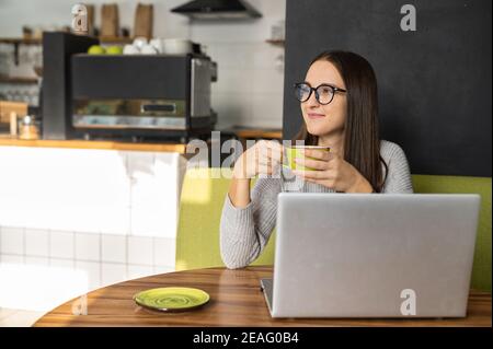 Nachdenkliche junge Frau macht mit einem Laptop eine Pause von der Online-Arbeit, sitzt am Schreibtisch in einem gemütlichen Café, hält eine Tasse mit Kaffee und schaut weg Stockfoto