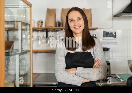 Selbstbewusste Geschäftsinhaberin, die mit gekreuzten Armen an der Theke eines Restaurants steht, blickt die Backwarenfrau im Vorfeld auf die Kamera und lächelt Stockfoto