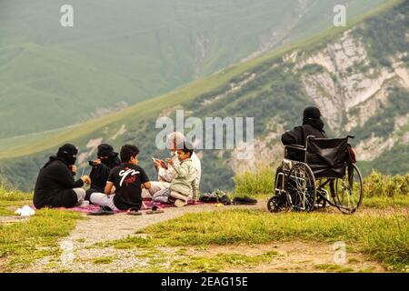 6-16-2019-Militärstraße, Georgien - Familie in islamischer Kleidung mit Mobiltelefonen - Frau im Rollstuhl sitzt auf der Klippe des Teufelstals im Kaukasus mou Stockfoto