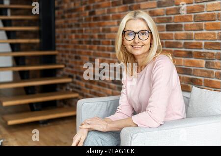 Porträt einer fröhlichen reifen blonden Frau auf dem Stuhl sitzen und lächeln vor einem Backstein Wand Hintergrund. Eine erfolgreiche Frau mittleren Alters in Brille Stockfoto