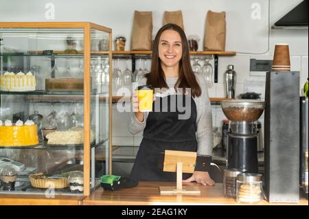 Freundliche und lächelnde Kellnerin im Schürze hält Papiertasse mit Take-Away-Kaffee in die Kamera. Fröhliche weibliche Bäckereibesitzerin bereitete heiße Getränke zu Stockfoto