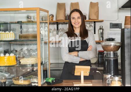 Lächelnde Kellnerin steht im Café und freut sich auf die Gäste. Weibliche Kleinunternehmerin mit gekreuzten Armen Stockfoto