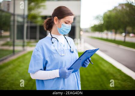 Weibliche UK NHS Arzt hält blaue Zwischenablage vor dem Krankenhaus stehen Klinik Straßeneingang, Frontlinie Schlüssel medizinischen Arbeiter studieren Patientenkarte Formular Stockfoto