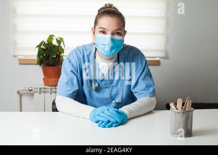 Junge weibliche kaukasische Allgemeinpraktizierende, die an ihrem Schreibtisch sitzt Büro, trägt blaue Uniform, Schutzhandschuhe & Gesichtsmaske, virtuelle Fernseher Stockfoto