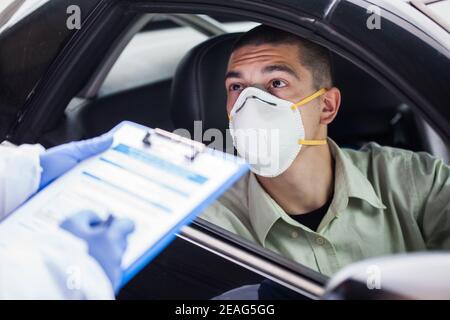 Männlicher Fahrer trägt eine schützende N95 Gesichtsmaske, die links sitzt Antriebsrad im britischen Drive-Thru COVID-19 Testzentrum, Beantwortung der Integritätsprüfung Nach oben Fragen Stockfoto