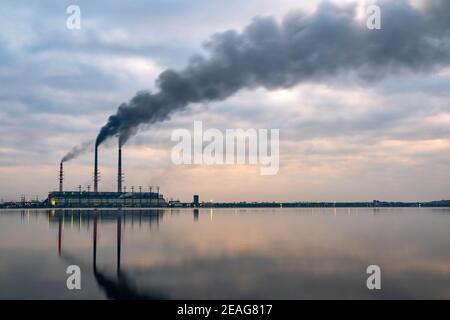Kohlekraftwerk hohe Rohre mit schwarzem Rauch, der nach oben die verschmutzende Atmosphäre mit den Reflexionen von ihm im Seewasser bewegt. Stockfoto