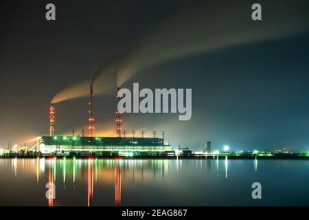 Hell erleuchtetes Kohlekraftwerk hohe Rohre mit schwarzem Rauch, der nach oben die verschmutzende Atmosphäre in der Nacht mit den Reflexen von ihm im Seewasser bewegt. Stockfoto