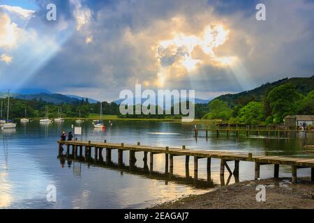 Sonnenstrahlen durch Wolken und eine hölzerne Anlegestelle am Lake Windermere im englischen Lake District gesehen bei Waterhead in Ambleside, Cumbria, Nordwestengland Stockfoto