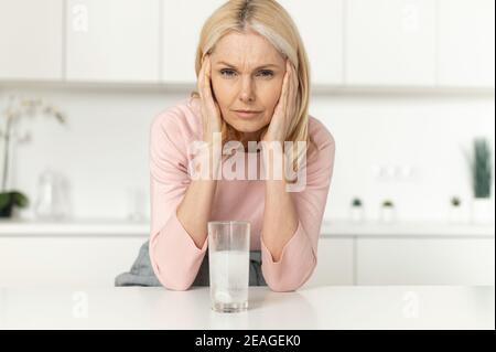 Eine müde reife blonde Frau, die in der modernen Küche steht, Tempel berührt, an Migräne oder schweren Kopfschmerzen leidet, Medizin Antibiotika nimmt Stockfoto