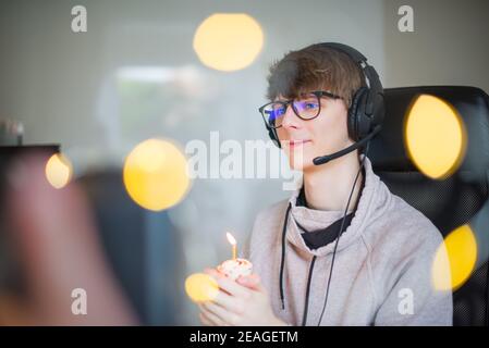 Junger Mann mit festlichem Cupcake mit brennender Kerze auf virtueller Geburtstagsparty während des Lockdown. Generation Z Mann feiert seinen Geburtstag allein auf Stockfoto