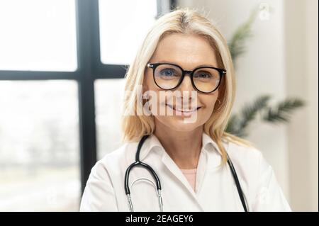 Headshot von intelligenten professionellen reifen weiblichen Arzt mit blonden Haaren, Tragen einer Brille, medizinische weiße Mantel und Stethoskop, Blick auf die Stockfoto