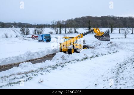 Ein Bagger räumt den Schnee für einen LKW, der 24 Stunden lang im Schneeverwehungsschnee stecken geblieben ist. Hoxne, Suffolk. Stockfoto