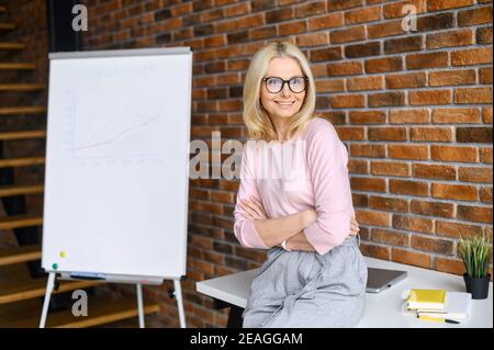 Reife Lehrerin, Coach in Brille mit blondem Haar, die Kamera anblickend, virtuelle Lektionen online per Zoom-Telefonkonferenz oder Geschäftsfrau Stockfoto