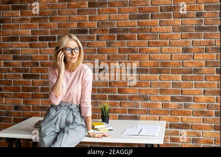 Elegant beschäftigt stilvolle reifen mittleren Alters blonde Geschäftsfrau, ceo in Gläsern sitzen am Rande des Büroschreibtisch in der modernen Arbeitsplatz und Stockfoto