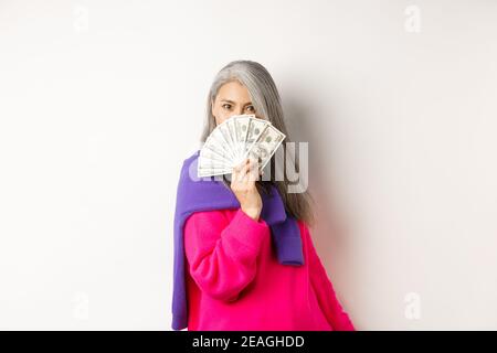 Einkaufskonzept. Reiche und modische koreanische ältere Frau versteckt Gesicht hinter Geld, Blick auf die Kamera, stehend über weißem Hintergrund Stockfoto