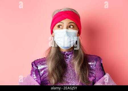 Covid-19, Virus und Modekonzept. Überrascht und erstaunt asiatische Großmutter in stilvollem Outfit und medizinische Maske, Blick links beeindruckt, rosa Stockfoto