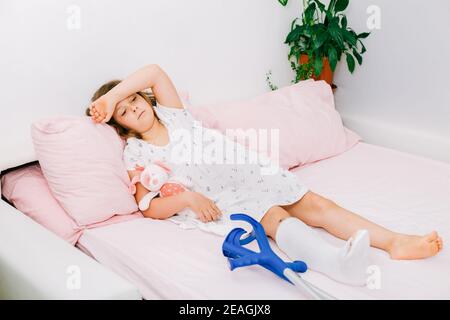 Ein trauriges Teenager-Mädchen liegt auf einem Bett mit einem Cast auf ihrem Bein, mit einem gebrochenen Knöchel und Krücken. Gebrochenes Bein in einem Kind.Nahaufnahme Stockfoto