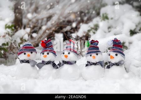 Fünf kleine niedliche Schneemänner in Hüten und Schals saßen im Winter im Schnee. Kaltes und eiskalt schnickendes Wetter. Weihnachten Urlaub Hintergrund Stockfoto