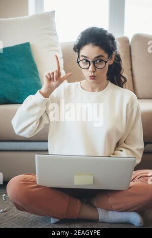 Lockig behaarte Frau ist gesturing die Idee Zeichen, während mit Online-Unterricht auf dem Boden am Laptop Stockfoto