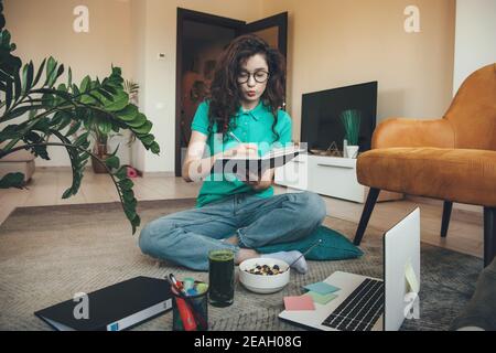 Kaukasische Mädchen mit lockigen Haaren ist mit Online-Unterricht auf Der Boden am Laptop beim Essen von Müsli und frischem Grün Saft Stockfoto