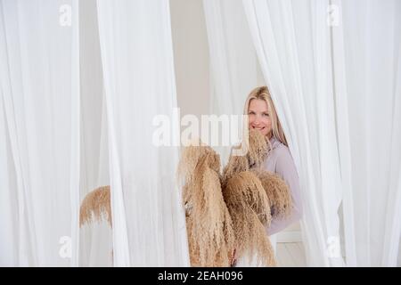 Junge blonde Frau in einem lila Hemd, weißes Kleid, hält in ihren Händen Strohkorb mit Pampas Gras zwischen den luftigen, Entwicklung Tüllstoffe. Minimali Stockfoto