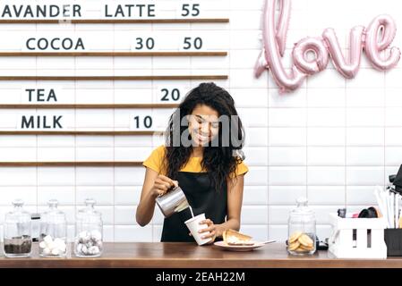 Glückliche afroamerikanische Barista in schwarzer Schürze macht Kaffee, während sie an der Theke steht, gießt sie Milch in ein Einwegkartonglas Kaffee mit freundlichem Lächeln Stockfoto