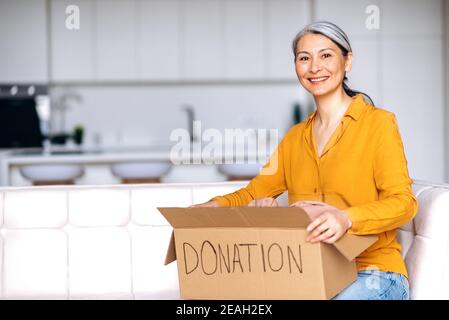 Spendenkonzept. Eine zufriedene Frau mittleren Alters mit grauem Haar, die zu Hause auf dem Sofa sitzt, hält eine Spendenbox mit voller Kleidung für die Wiederverwendung oder das Recycling alter Kleidung. Wohltätigkeitsorganisation. Hilfe für arme und bedürftige Menschen Stockfoto