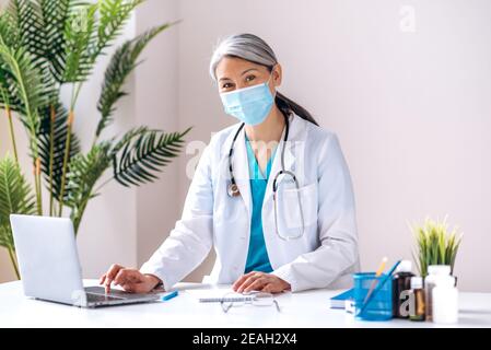 Porträt einer Ärztin mittleren Alters mit grauem Haar in Laborkittel und medizinischer Maske sieht direkt auf die Kamera am Arbeitsplatz in der Klinik. Ärztliche Beratung, Gesundheitskonzept Stockfoto