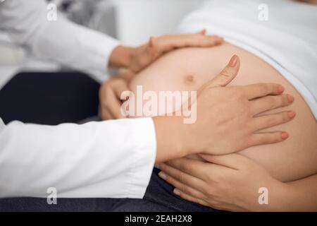 Nahaufnahme einer schwangeren Frau, die auf der Couch liegt, während die Ärztin ihren Bauch manuell untersucht. Gynäkologe tun Palpation des Patienten im modernen Krankenhaus Stockfoto
