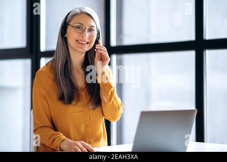 Freundliche asiatische reife grauhaarige Geschäftsfrau oder Tutor, in lässiger stilvoller Kleidung, Headset und Brille arbeitet online, oder führen Abstand Unterricht, mit Laptop am Arbeitsplatz, schaut auf die Stockfoto